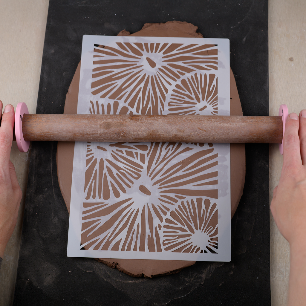 Person using a rolling pin with a decorative stencil to imprint patterns on clay.